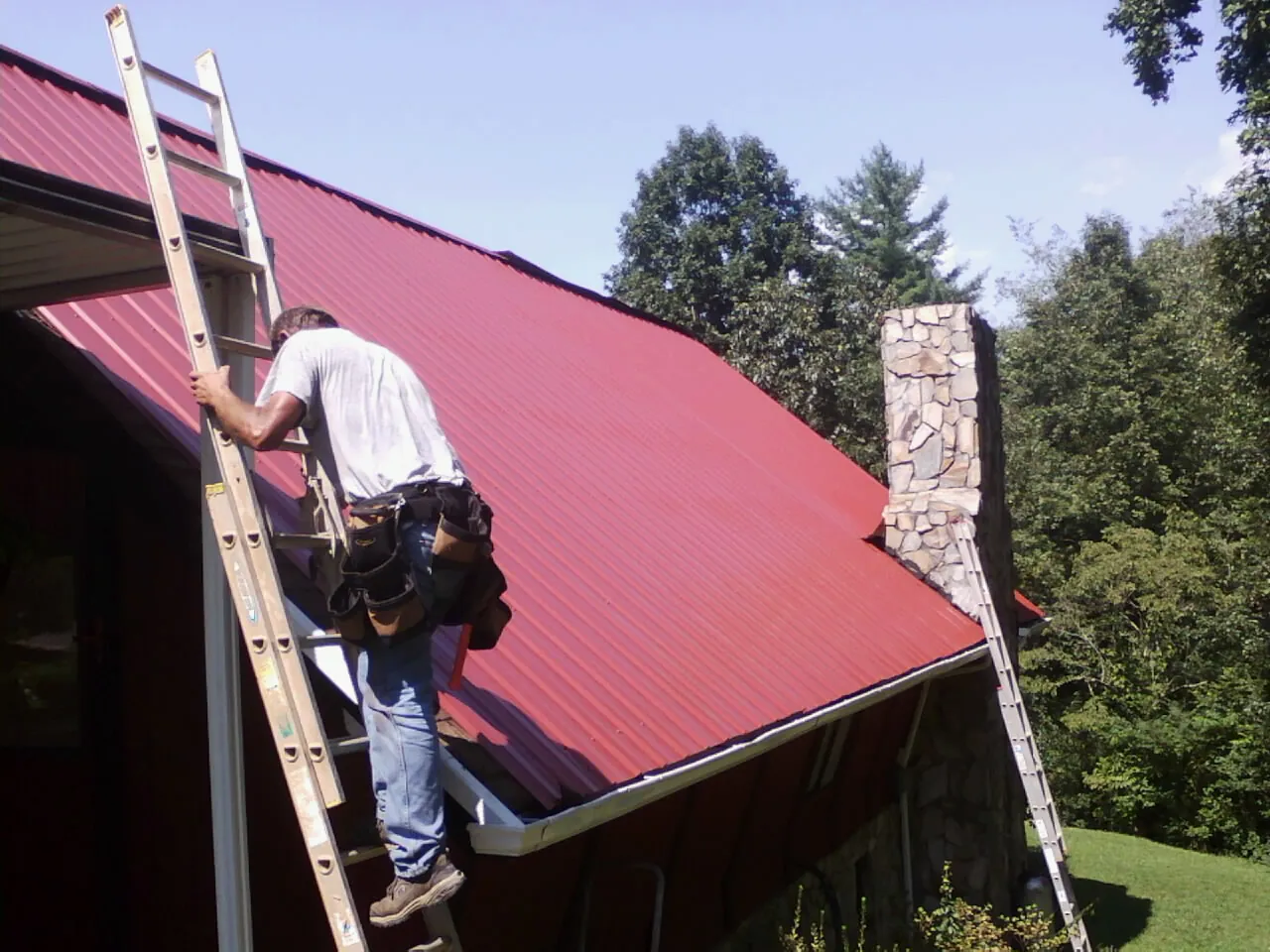 Roofer on a ladder beside a red metal roof and stone chimney — Soffit & Fascia Repair by Roofing Brothers in Shakopee
